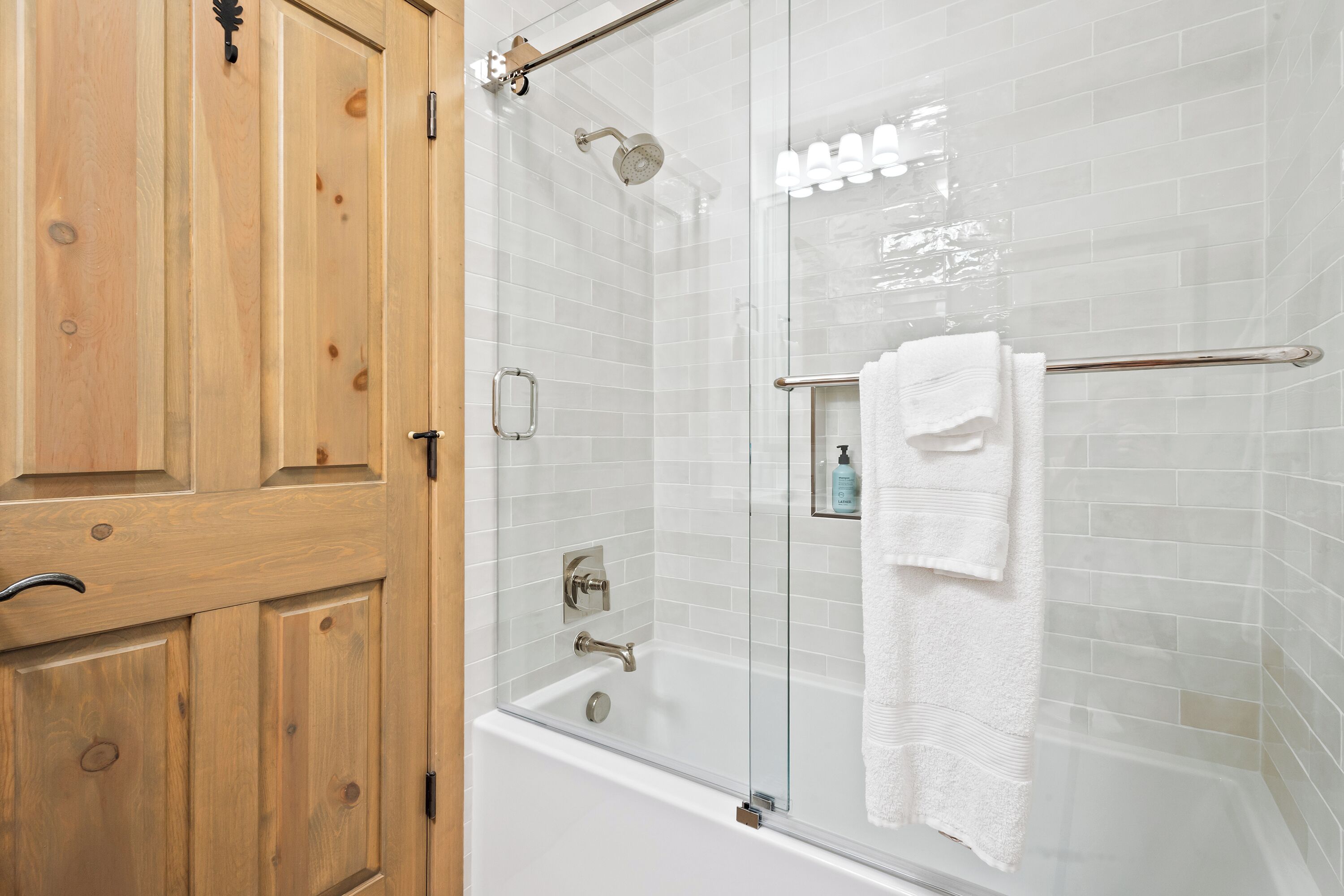 A glass-enclosed shower area with a white tiled wall, a wooden door, and a towel rack with a white towel.