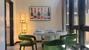 A dining area with a round table set for four, green chairs, a chandelier, and a wall-mounted artwork of bottles.