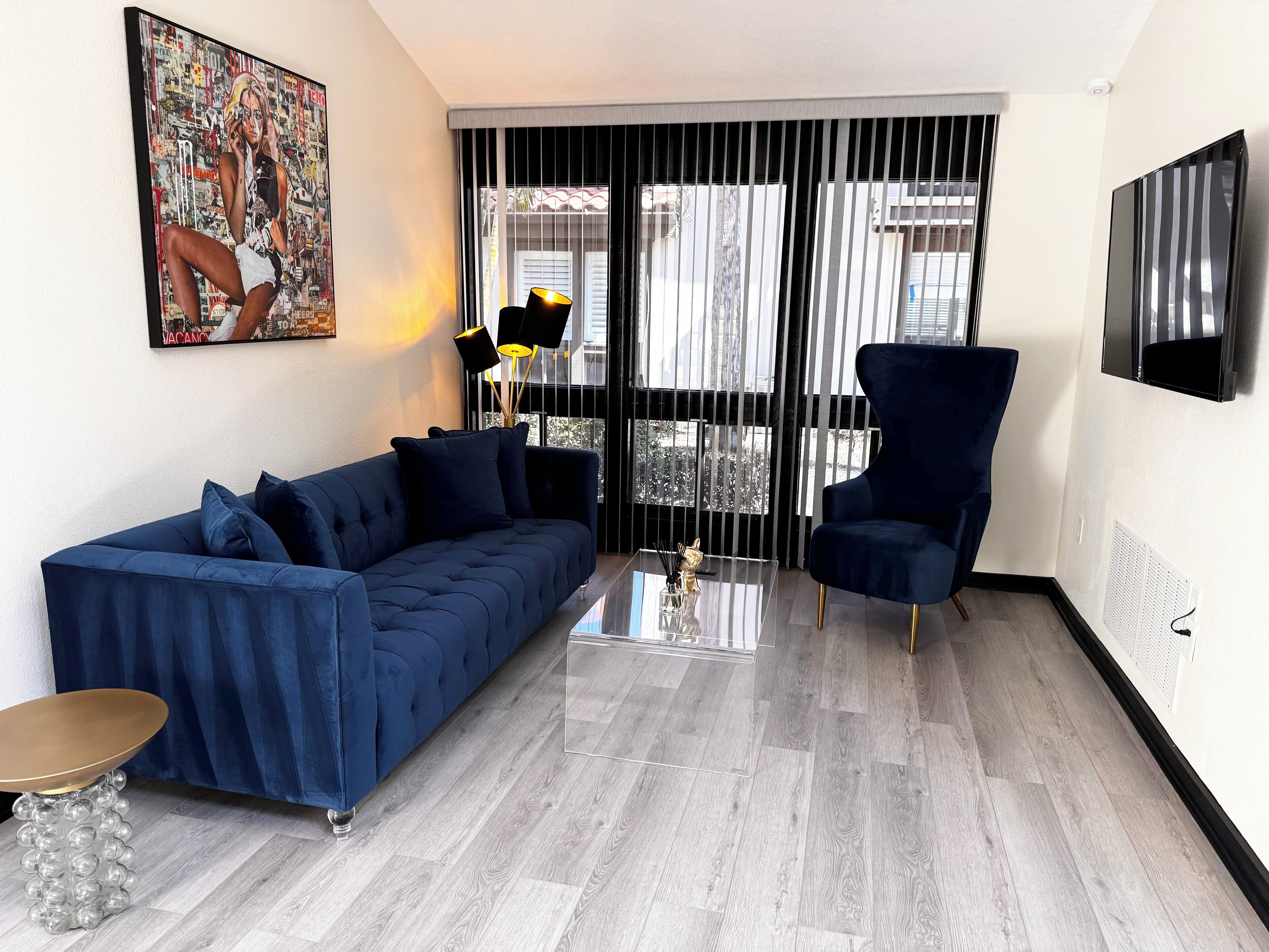 A modern living room with a blue sofa, a matching armchair, a glass coffee table, and a painting on the wall.