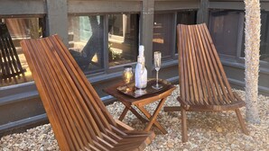 Two wooden lounge chairs with a tray table, a bottle, and a glass on a pebble-covered ground, in front of a building with large windows.