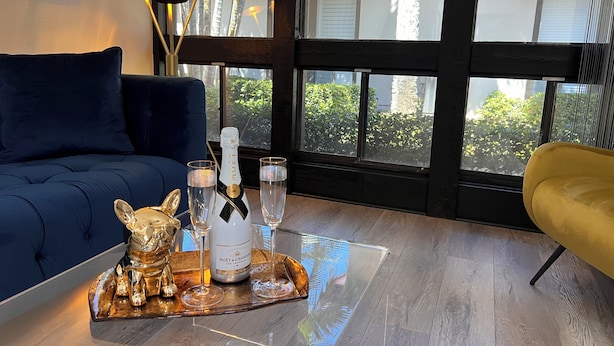 A room with a blue sofa, a yellow armchair, a glass table with a bottle of champagne and glasses, and a large window with a view of greenery.