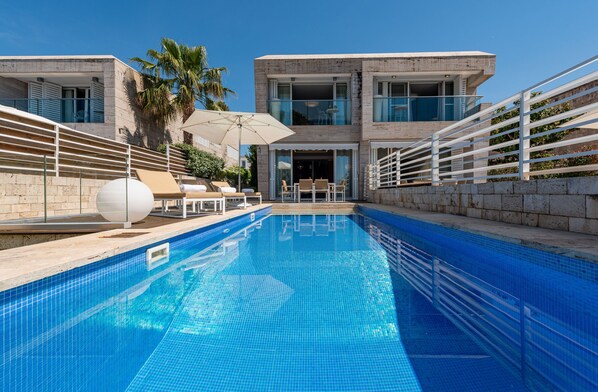 Ein moderner Poolbereich mit klarem, blauem Pool, Liegestühlen und einem zweistöckigen Gebäude mit großen Fenstern und einem Balkon.