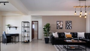 A modern living room with a black leather sofa, a coffee table, and a bookshelf. There is a door leading to a bathroom and a plant in the corner. - Zest City Residence by Nomads (Paphos)
