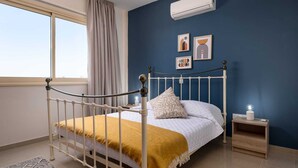 A bedroom with a white metal bed frame, a white bedspread, a mustard-colored throw blanket, a bedside table with a lamp, and an air conditioning unit on the wall. - Zest City Residence by Nomads (Paphos)