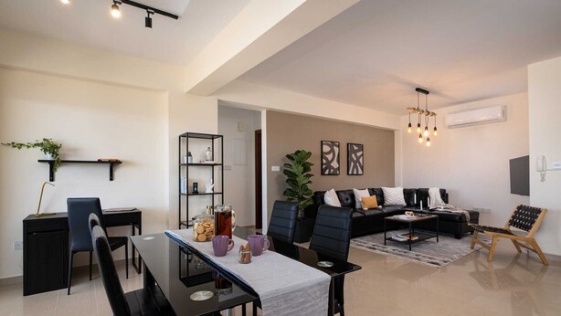 A modern living room with a black leather sofa, a dining table with chairs, and a coffee table. There is a bookshelf, a potted plant, and a hanging light fixture. - Zest City Residence by Nomads (Paphos)
