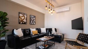A modern living room with a black leather sofa, a wooden coffee table, and a black and wooden chair. There are abstract paintings on the wall, a ceiling light fixture, and an air conditioning unit. - Zest City Residence by Nomads (Paphos)