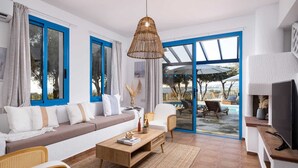 Ett modernt vardagsrum med en soffa, ett soffbord och stolar. Det finns en tv på ett tv-bord, en hängande lampa och en utsikt över ett utomhuspoolområde genom glasdörrar.