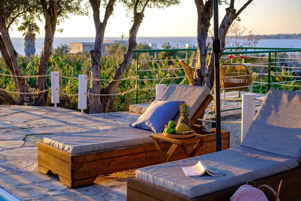 A beachside lounge area with wooden loungers, a small table with fruits, and a view of the sea.
