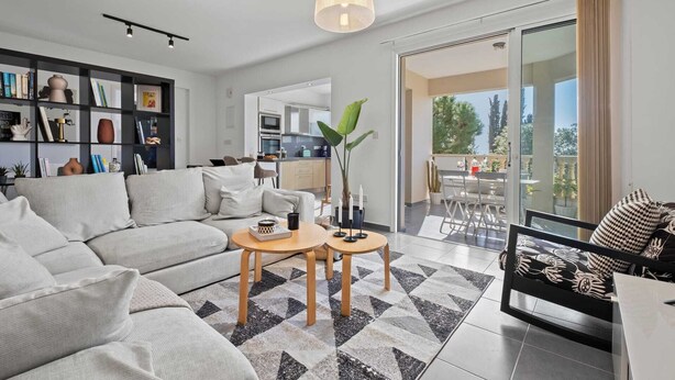 Ein modernes Wohnzimmer mit einer Couch, einem Couchtisch und Sesseln, eine Küche im Hintergrund und ein Balkon mit Gartenmöbeln.