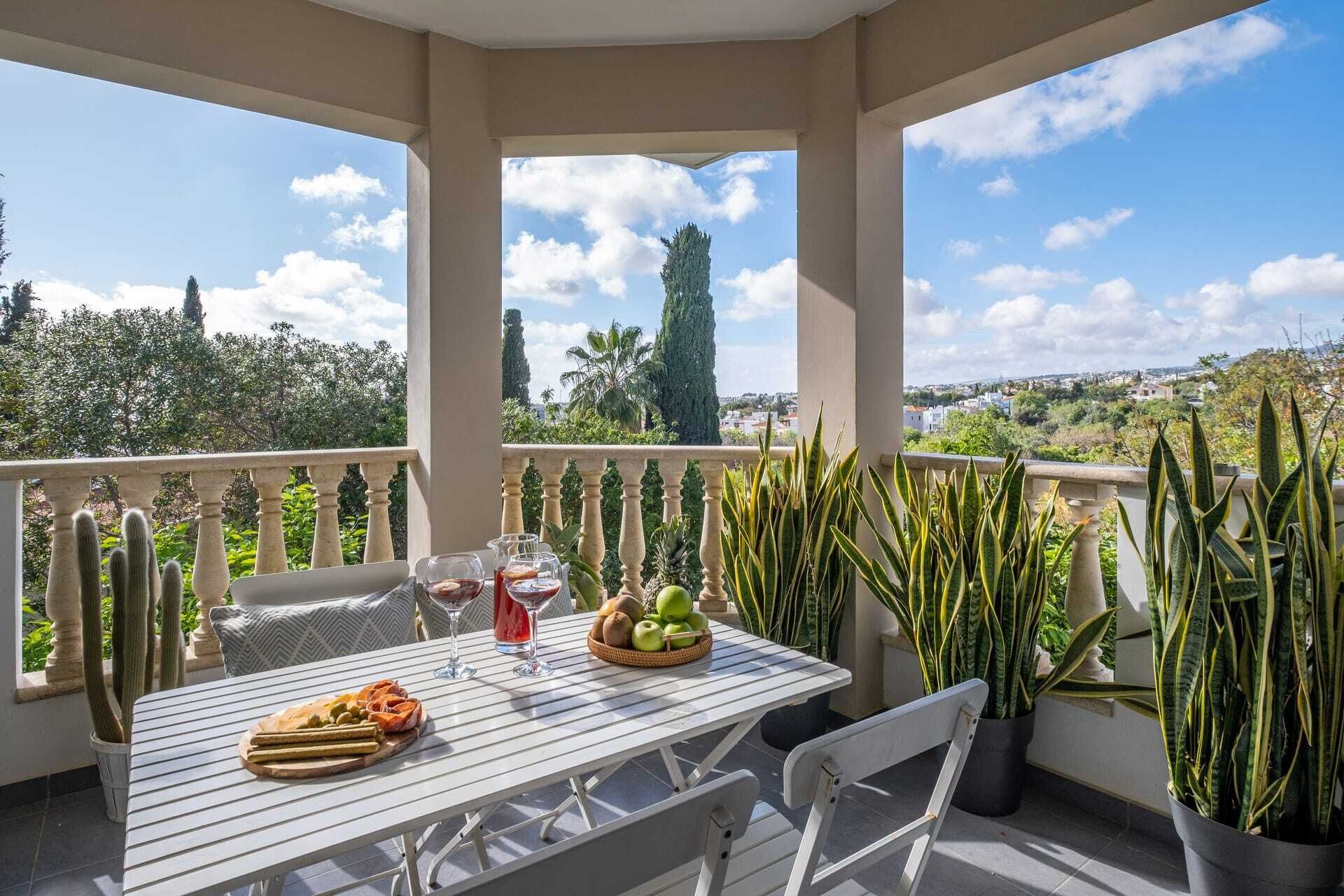 Un balcón con mesa lista para comer, rodeado de plantas en macetas y con vistas panorámicas.