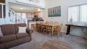 A spacious living room with a brown sofa, a dining area with wooden chairs, a kitchen in the background, and a staircase leading to an upper level. - 8280 The peak- Hot tub, ski in/ ski out, Pool table, Golf course, fire pit (Clymer)