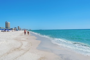 Een strand met een heldere lucht, enkele mensen die wandelen en een stadsachtergrond.