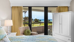 A hotel room with a bed, a chair, a nightstand, a lamp, and a wardrobe. The room has a large window with a view of the ocean and palm trees. - 🚢Steps to Private Beach-Gulf Views-2 Pools with Waterfall-1BR Edgewater #303🚢 (Miramar Beach)