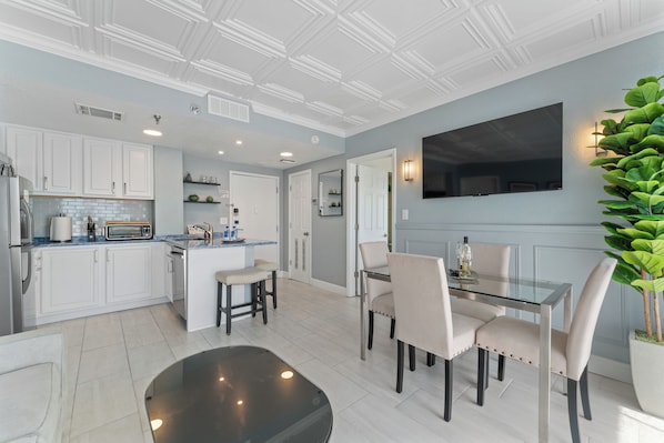 A modern kitchen with white cabinetry, a central island, and a dining area with a glass table and chairs. - Terrace Suite with Epic Fireworks View (Orlando)