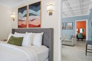 Habitación de hotel con una cama, dos cuadros en la pared, una mesita de noche y una puerta que da a otra habitación.