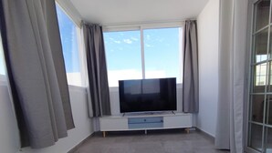 En moderne stue med et stort fladskærms-tv på et hvidt stativ, et unikt, buet sofabord og store vinduer med gardiner.