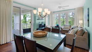 A spacious dining area with a glass table, chairs, and a chandelier. There is a sofa and a television in the background. - Elation Escapes (Miramar Beach)