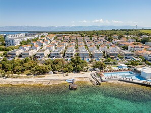 An aerial view of a coastal residential area with multiple buildings, clear turquoise waters, and a distant mountain range.