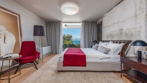 A modern bedroom with a large bed, a red chair, a desk, and a view of the sea. - Villa Oleander Zadarvillas (Zadar)