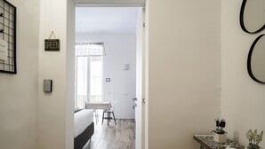 Ein schmaler Flur führt zu einem Schlafzimmer mit Bett, Nachttisch und Stuhl. Auf der rechten Seite steht ein kleiner Tisch mit Topfpflanzen.