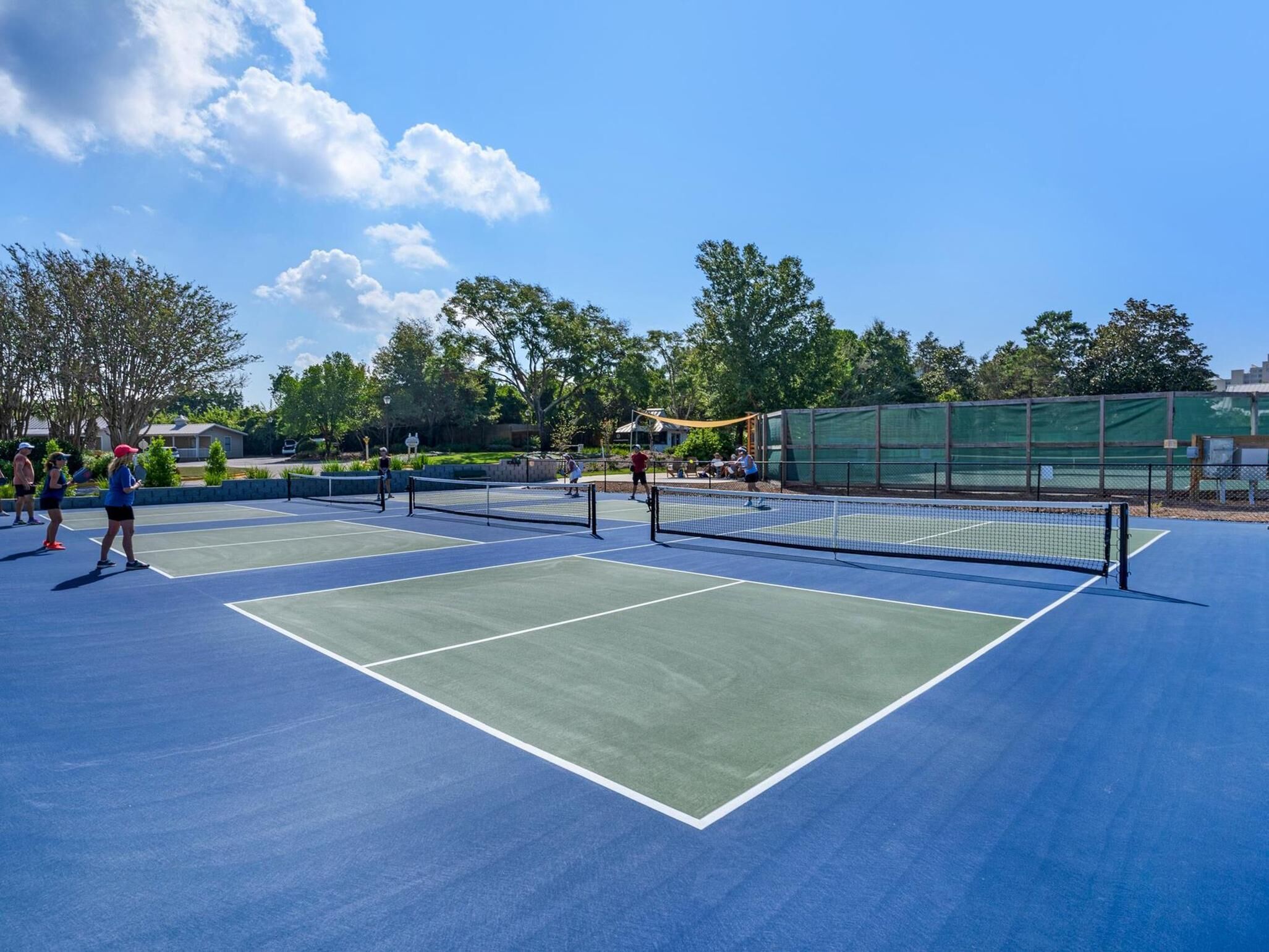 An outdoor tennis court with multiple players, surrounded by trees and a clear sky.