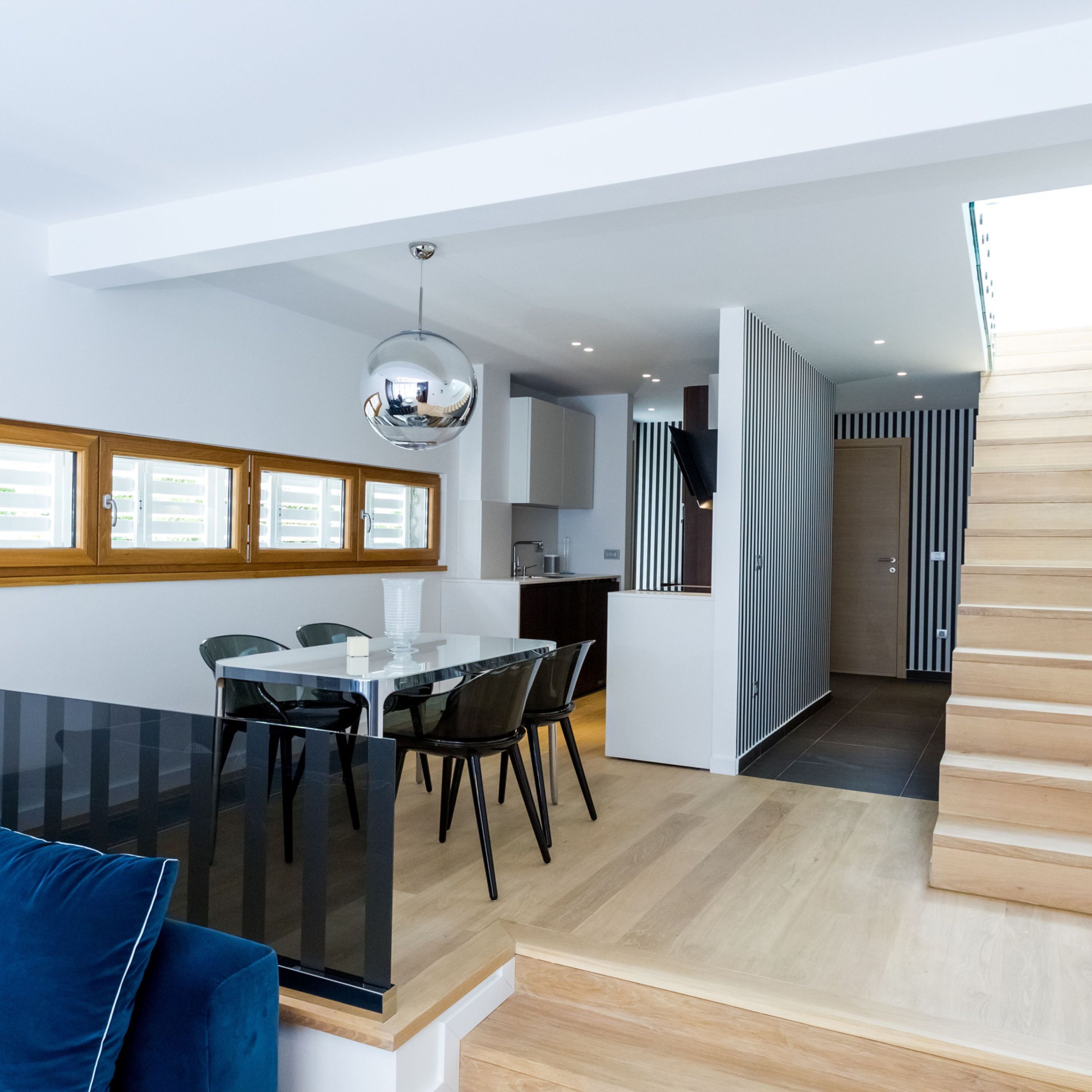 A modern kitchen with a central island, a dining area with a glass table and chairs, and a staircase leading to an upper level.