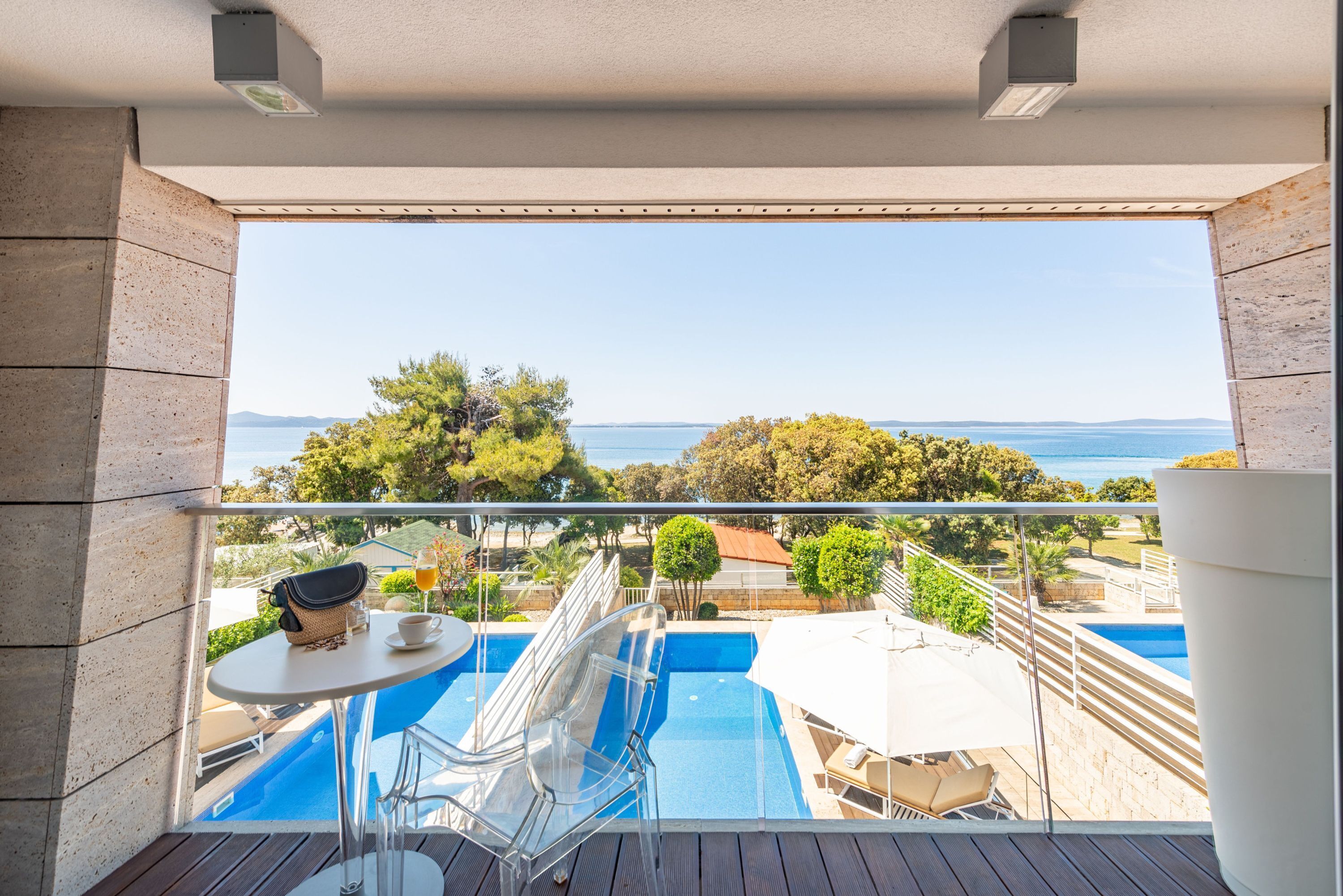 A balcony with a clear view of a swimming pool, lounge chairs, and a table with a bag and a glass of drink.