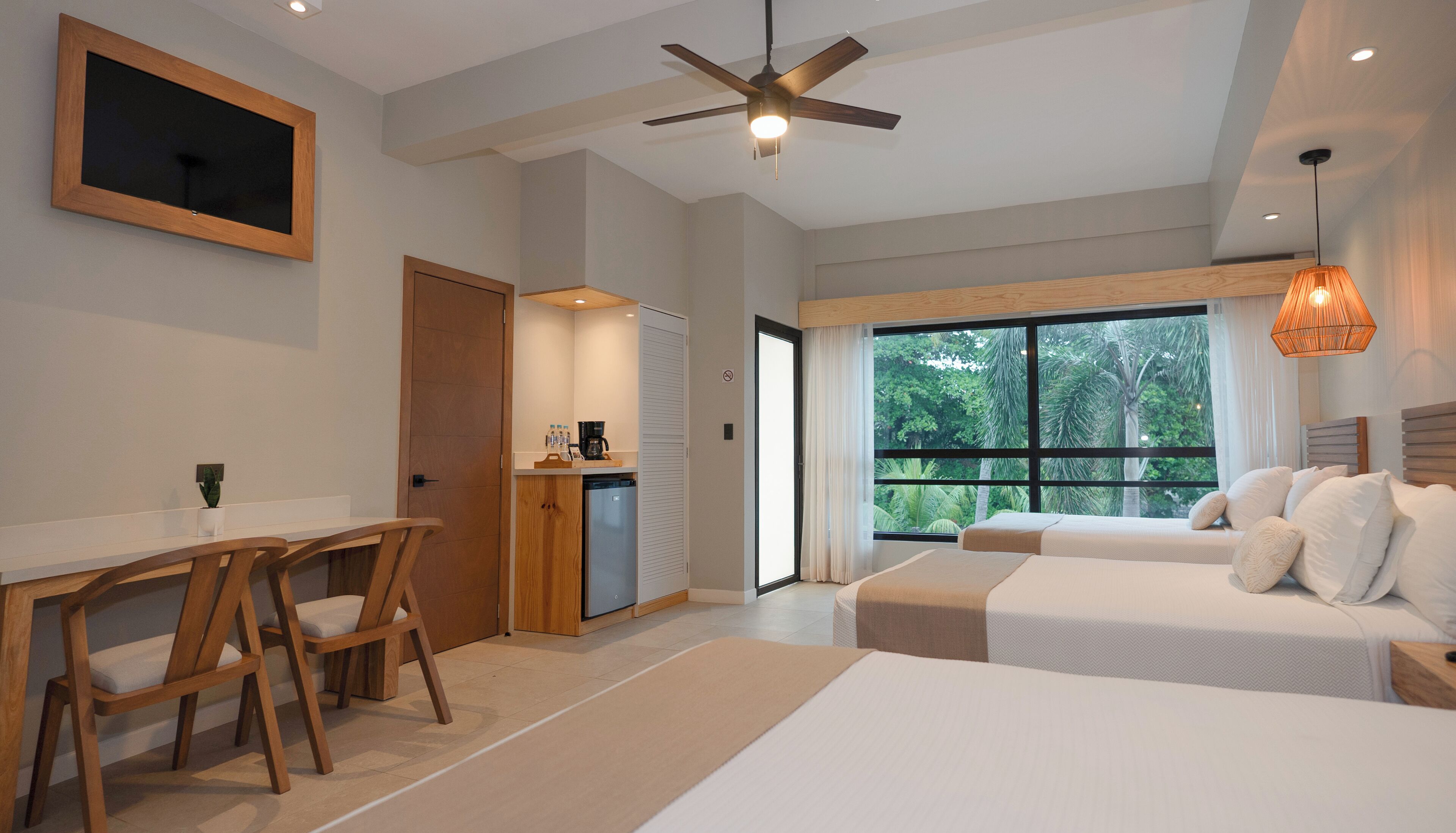 A modern hotel room with a large bed, a desk with a chair, a coffee machine, and a view of greenery through the window.