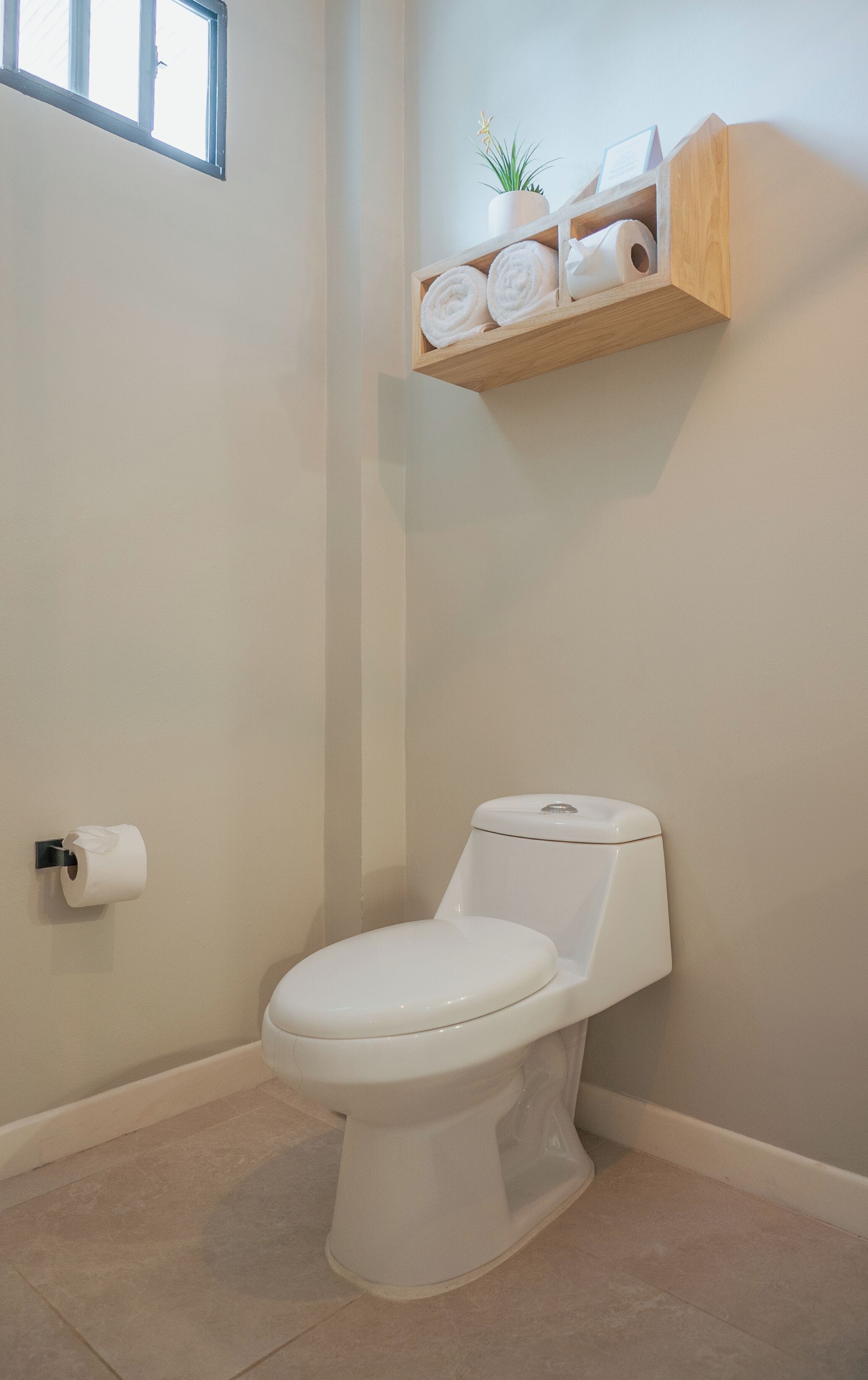 A white toilet with a white seat and lid, a toilet paper roll on the wall, and a wooden shelf with towels and a small plant.