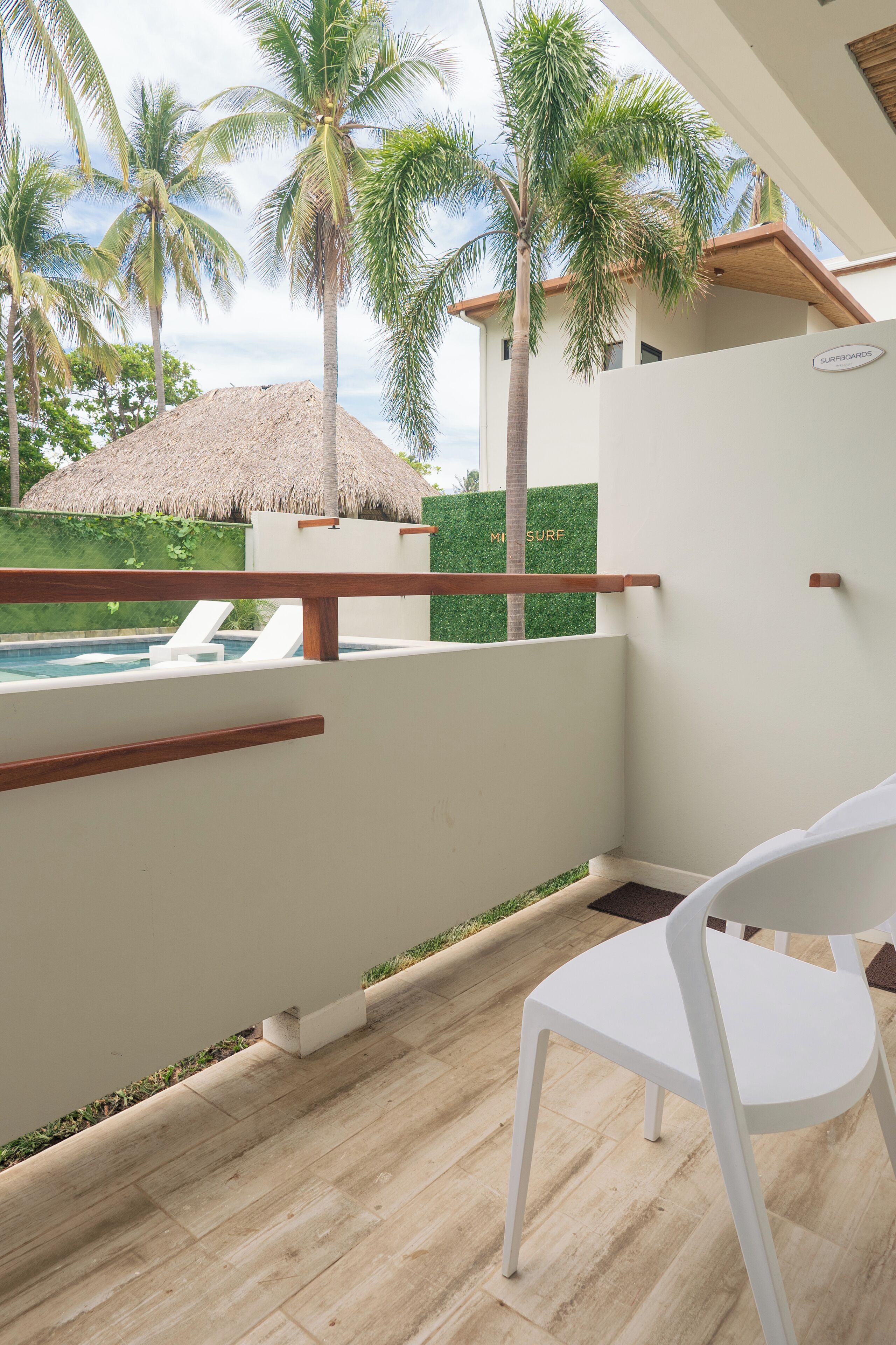 Uma área junto à piscina com uma espreguiçadeira branca, um guarda-sol de palha e palmeiras ao fundo.