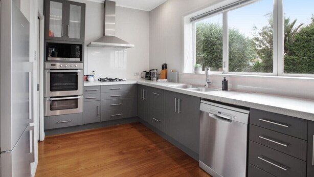 A modern kitchen with grey cabinets, stainless steel appliances, and a large window offering a view of greenery. - Prosper Point Home with BBQ, Patio and Parking (Auckland West)