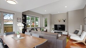 A modern living room with a dining area, grey sofas, a TV, and a fireplace. - Prosper Point Home with BBQ, Patio and Parking (Auckland West)