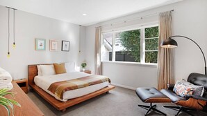 A modern bedroom with a wooden bed, a bedside table, a chair, a desk, and a window with curtains. - Prosper Point Home with BBQ, Patio and Parking (Auckland West)