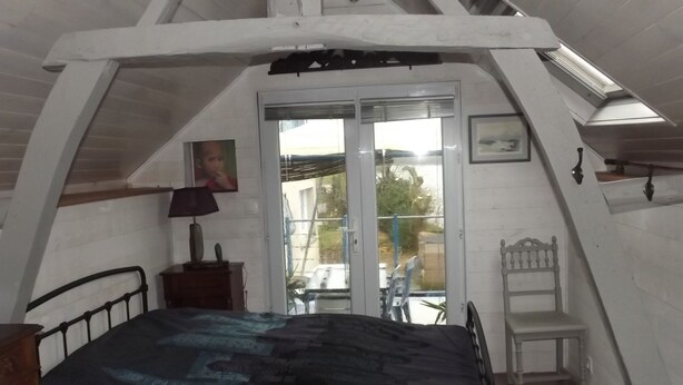 A bedroom with a sloped ceiling, a bed, a chair, a lamp, and a door leading outside. - Le Gîte des Pêcheurs au Calme à 5 mn de Terra Botanica (Cantenay-Épinard)