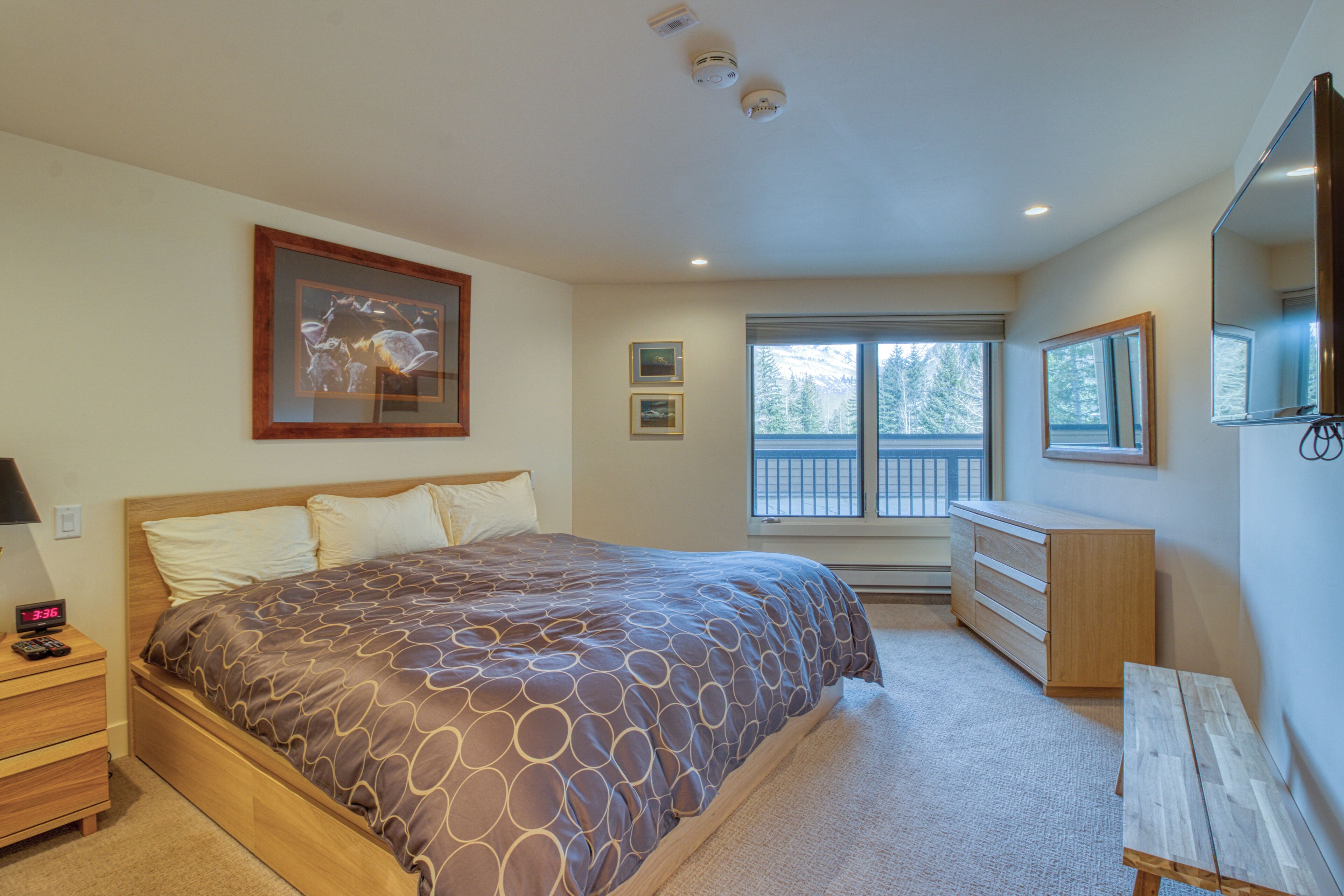 A bedroom with a large bed, a dresser, a nightstand, a window with a view of trees, and a ceiling with recessed lighting.