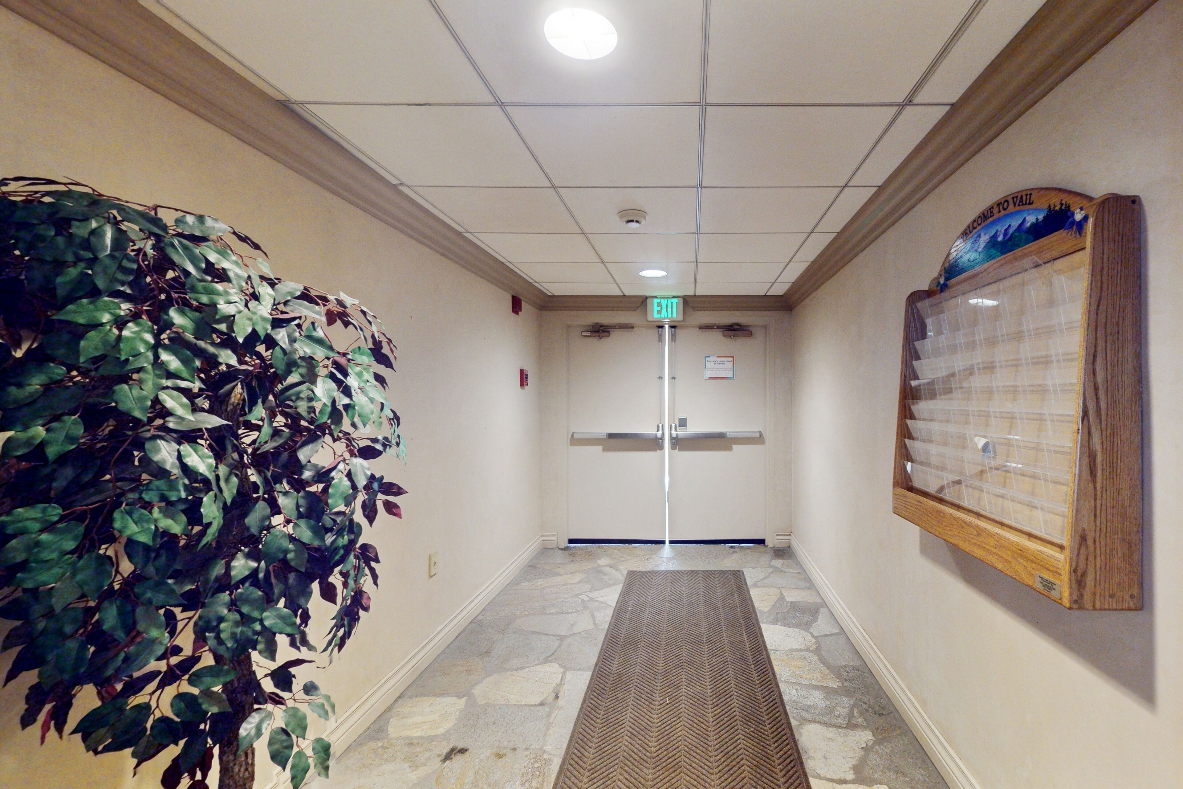 A hallway with a stone-patterned floor, an exit sign, a welcome sign, and a wall-mounted bulletin board.