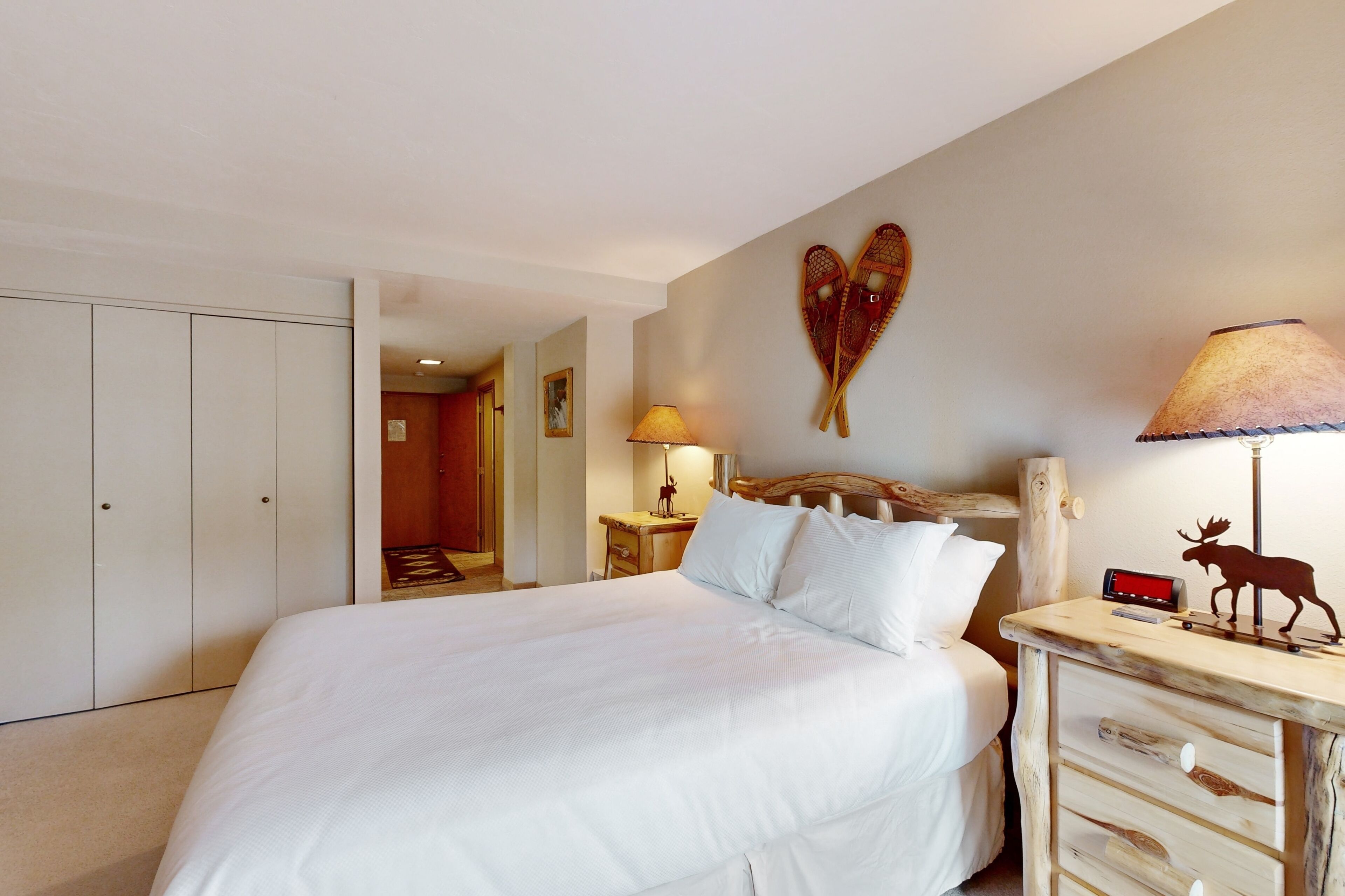 A hotel room with a large bed, a wooden nightstand, a lamp, a moose decoration, and a heart-shaped wall art.