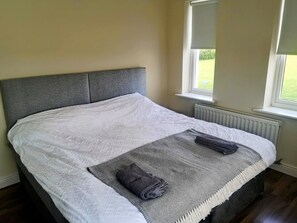 A neatly made bed with a grey headboard and a white quilt, a grey towel on the bed, and a window with a view of greenery. - 2 Bedroom House Near Athy (Barrowhouse)