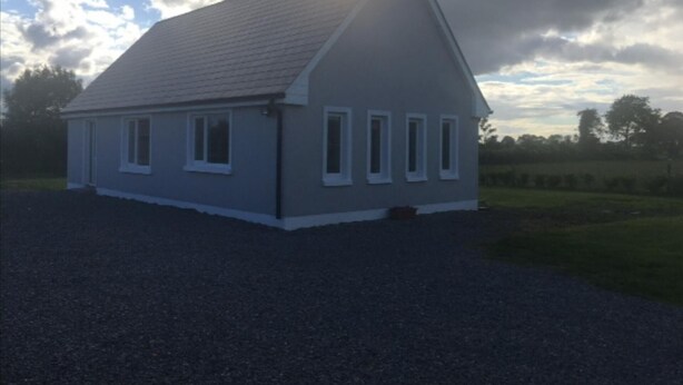A small, single-story house with a sloped roof, a gravel driveway, and a rural setting with trees and open fields in the background. - 2 Bedroom House Near Athy (Barrowhouse)