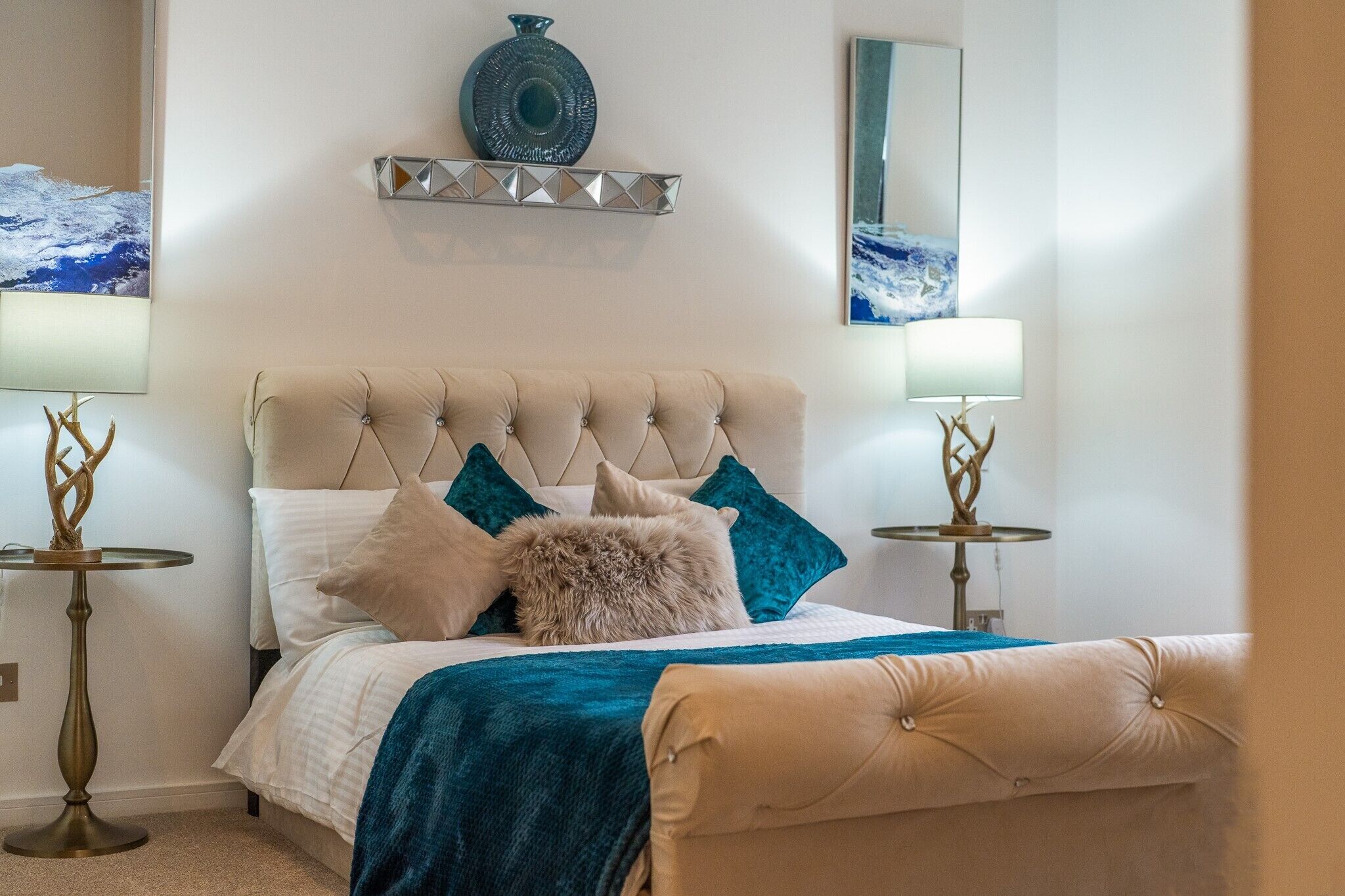 Un dormitorio ordenado con un cabecero capitonado, una cama con una manta color turquesa y dos mesitas de noche con lámparas.