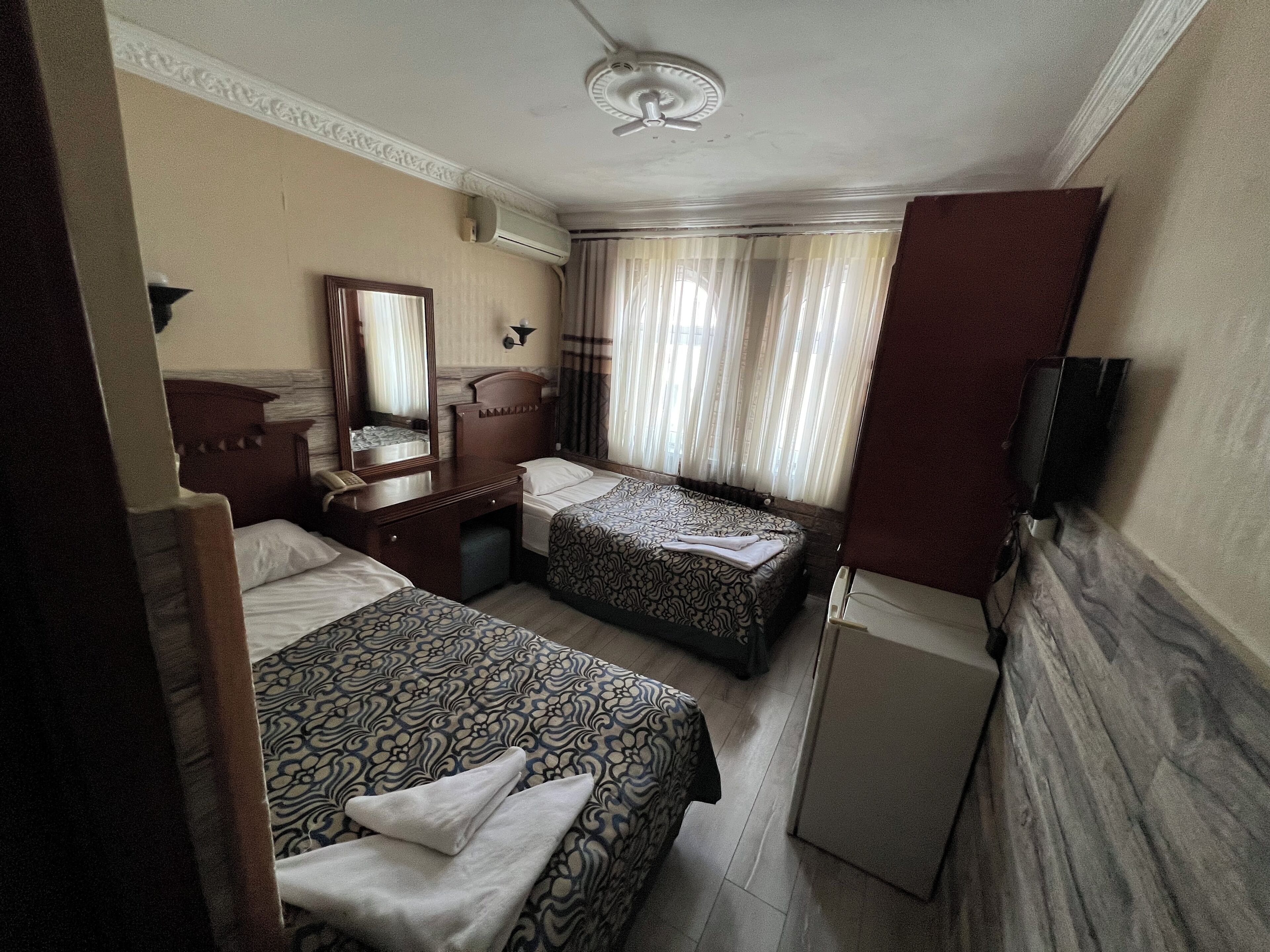Habitación de hotel con dos camas, un escritorio con teléfono, un espejo, un televisor de pantalla plana y un ventilador de techo.