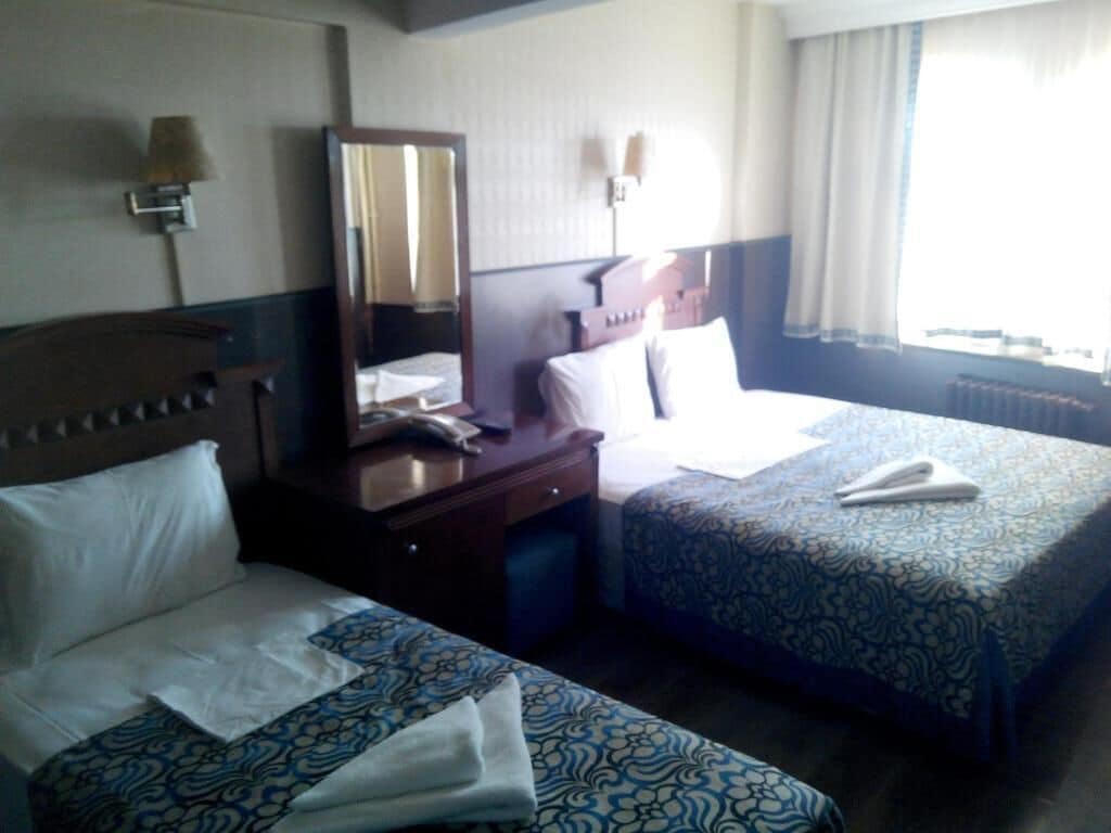 A hotel room with two beds, a bedside table, a mirror, and a window with curtains.