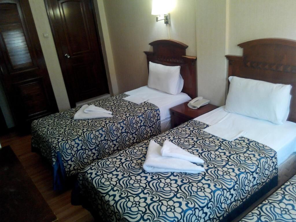 A hotel room with two beds, each with patterned bedding and a wooden headboard. There is a nightstand with a telephone and a folded towel on each bed.