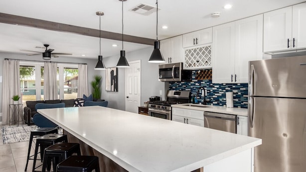 Une cuisine moderne avec des meubles blancs, un grand îlot central, des appareils en acier inoxydable et des luminaires suspendus.