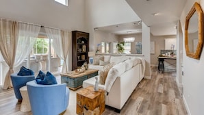A spacious living room with a large white sofa, blue armchairs, and a wooden coffee table. A dining area is visible in the background with a table and chairs. - Pickle Ball, Heated Pool, Spa, Outdoor Bar w/Swings, Game Room, Bocce, & Putt-Putt! (Scottsdale)