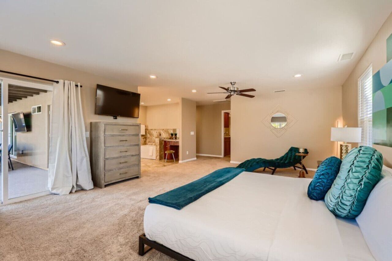A bedroom with a bed, a dresser, a TV, a ceiling fan, and a view of a kitchen and bathroom.