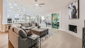 A modern living room with a grey sectional sofa, a coffee table, and a fireplace. There is a kitchen in the background with white cabinets and a dining area with a table and chairs. - Resort Style Estate, 2 Houses, Putt-Putt Course, B-ball Court, Heated Pool (Scottsdale)