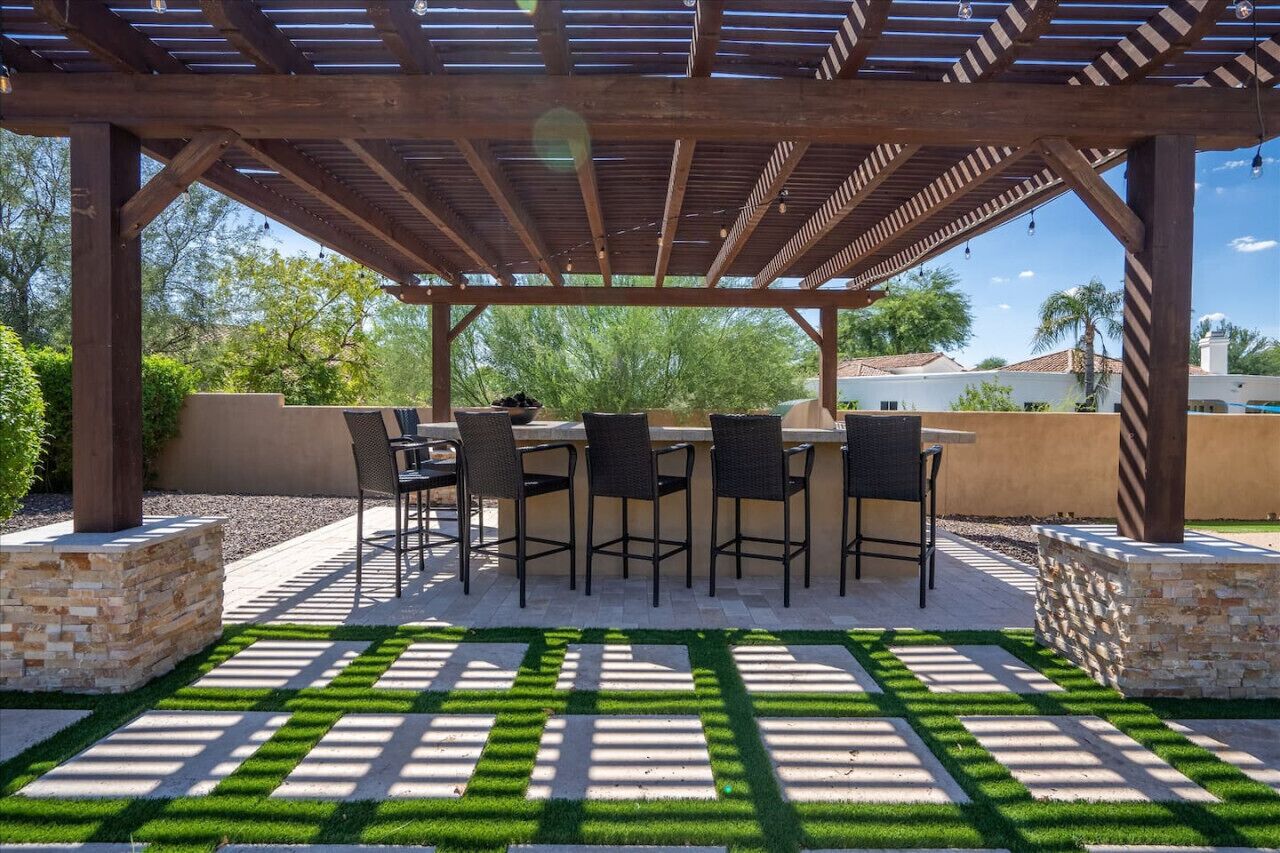 Un patio exterior con una pérgola, una mesa con sillas y jardineras de piedra.