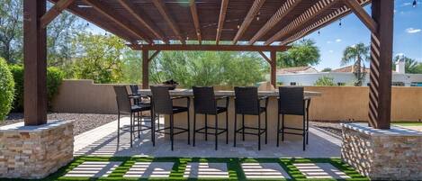 Un patio exterior con una pérgola, una mesa con sillas y jardineras de piedra.