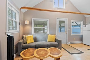 A living room with a gray sofa, wooden coffee tables, and a kitchen area with a refrigerator. - Tiger Lily Cabin: Sunset Shores Kangaroo Lakefront (Baileys Harbor)